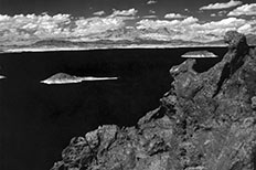 Textured mountain in lower right, lake with water appearing black has two islands, cumulus clouds in distance.