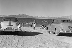 Car at right and left, people on beach and in water behind.