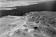 Aerial view of large hills at lower right, cars in parking area to lower left, lake with many boats upper area.