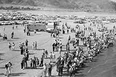 Beach crowded with cars and people, mountains in distance.