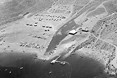 Aerial view, lake is at bottom of frame, paved launch ramp at center divides areas of parked cars and buildings.