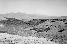 Large curved desert valley between rugged large hills, mountains in distance. 