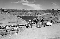 Many cars parked along a road at left, leading to a lake, two camping trailers in the foreground.