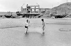 Two men in water using a fishing net, dam under construction in distance.