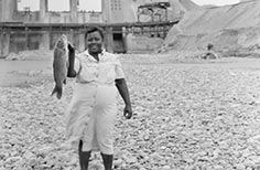 A smiling woman holds a fish with one hand, dam under construction behind.