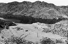 Camping trailers, vans, and small structures near a dirt road leading to a lake with a mountain surrounding it.