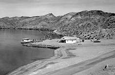 Small dock on a lake with a structure and two cars nearby along a dirt road in the foreground.
