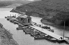 17 boats tied to boat dock with a building, the dock is in middle of a small cove.