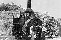 Two men stand beside a large steam powered tractor.