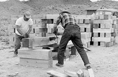 Two men work together with hand saw to cut large wooden beam, cut beams stacked in background.