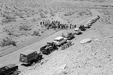 Dirt road at angle divides desert landscape, row of cars to right, crowd of people to left.