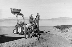Man digs trench with back hoe, another man smooths trench with rake, lake and mountains in distance.