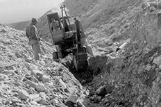 Man operating a trenching machine between rock formations as another man watches.