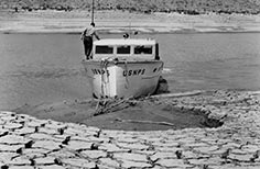 Man on a boat on a lake with dried up and cracked mud on nearby beach.