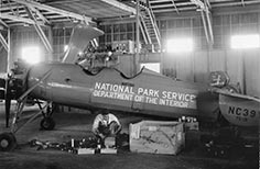 National Park Service autogyro in a hangar with man kneeling down next to a crate and machine parts.