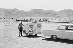 Man in uniform stands next to cart full of trash being pulled by a car.