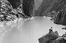 A river surrounded by towering cliffs, a man very small in photo, sits on rock formation at edge of water at lower right.