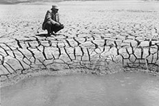 Man in hat squats at edge of pool of water surrounded by dry cracked mud.
