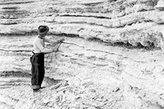 Man wearing a hat places his hand on large sedimentary rocks as he holds a hammer in the other. 

