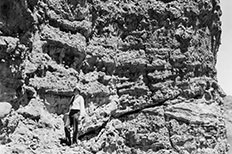 Man in white shirt, tie and hat stands in front of a textured cliff.