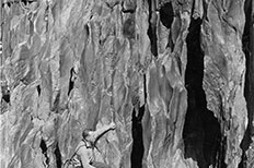 Man in lower left sits on small outcropping of rock protruding from large cliff face.