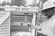 Man with thermometer his hand in front of a wooden enclosure labeled US NPS 