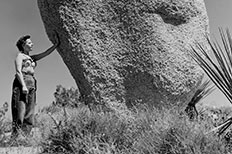 Woman stands next to large balanced boulder, her  her hand is on it, surrounded by desert plants.