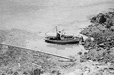 Log boom filled with driftwood in the lower left corner with a boat floating in a body of water near the desert shoreline. 
