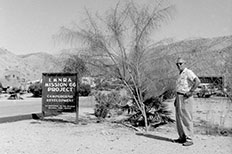 Man in sunglasses poses next to a tall Palo Verde tree and campground development sign with campers and mountains in the distance. 