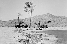 Desert landscape with a rocky trench-like pathway containing planted trees and shrubs with a parked pick-up truck and man operating a backhoe loader in the distance.