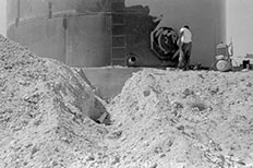 Man painting a large steel water tank with a ladder attached, desert landscape with a deep trench in the middle and a vertical pipe sticking out.