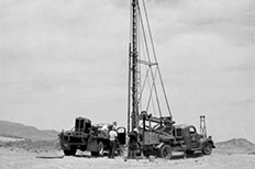 Two men stand in-between two trucks, one truck has large drilling rig attached.

