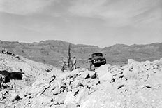 Two men wearing hats operate a machine with a parked pick-up truck to the right surrounded by rock formations and mountains in the distance.