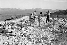 Four men stand among rubble of rocks, mountains in the distance.

