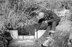 Man wearing a hat measures water level of small stream.