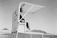 Woman sits on covered lifeguard stand as she looks at a large body of water while two people float on a dock with mountains in the distance.