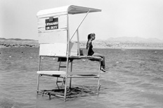 Woman sits on covered lifeguard stand as she looks at a large body of water and distant mountains.