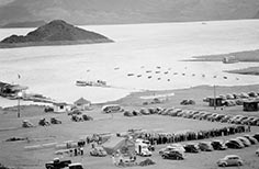 Crowds of people wait in a line in a dirt parking lot next to a lake.