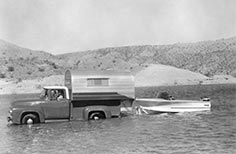 Man in truck with camper shell launches boat, rolling desert hills in background.