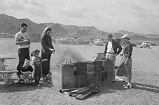 A woman roasts hot dog on stick at a brick barbeque, two children and three adults with her.