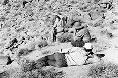 Two men sit on rocky desert landscape, another laying down with back to camera.