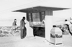 Girl in pigtails sits on small stone wall by man looking at National Park Service bulletin board as people stand nearby.