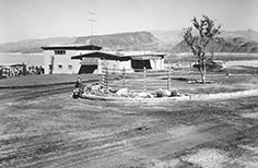 Man sits on curb at dirt road with small building nearby.