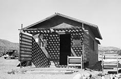 Small building with two wooden beds outside to the right and a car back towards the left.