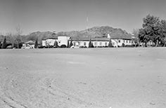 Dirt road leading up to large building with cars parked nearby it.