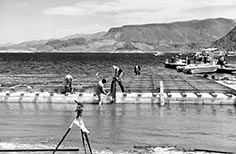 Several people constructing a dock on a lake with a surveyor's instrument in the foreground.