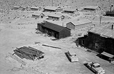 Several small buildings with a stack of wood and two cars nearby.