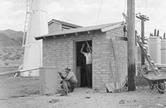 Small stone building with man standing in doorway and another man out front kneeling down and painting.