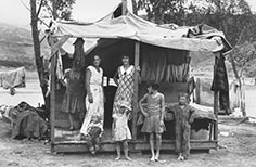 Women with children stand on porch of small cabin.