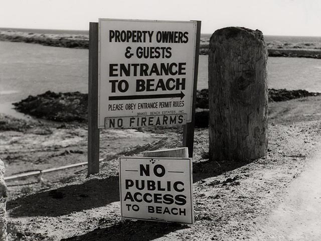 All photos in this gallery are black and white. This one shows a beach adjacent a road with signs that say “Property Owners & Guests Entrance to Beach, please obey entrance permit rules” and “No Public Access to Beach.”